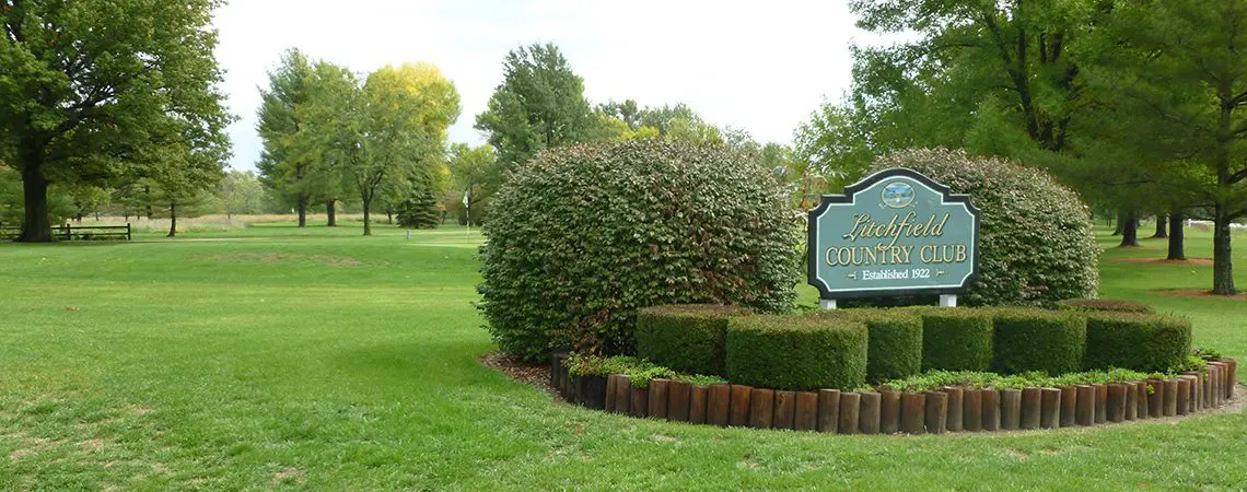 A sign in the middle of a park with bushes around it.